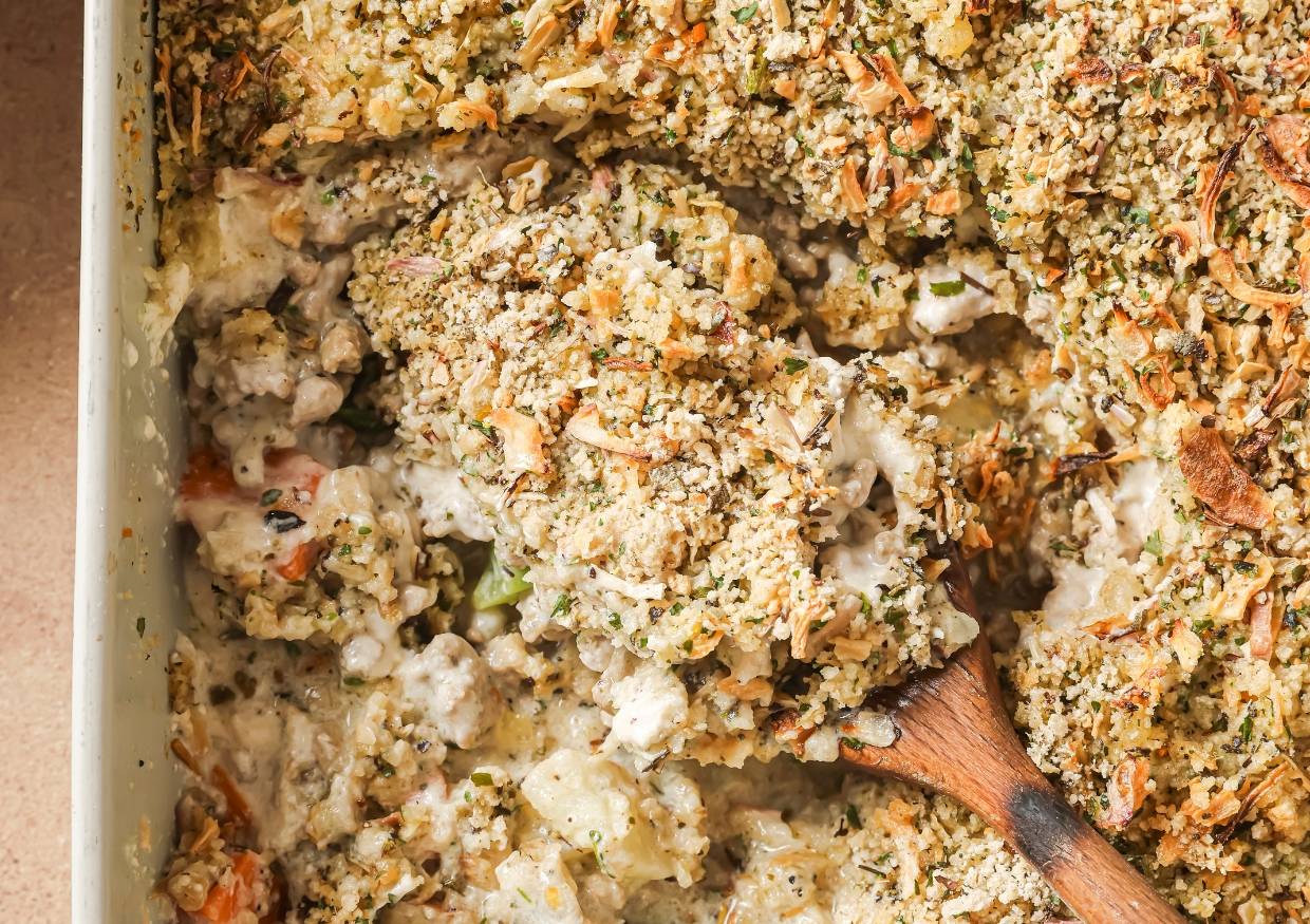 A close-up of a creamy casserole with a golden, crunchy breadcrumb topping, herbs, and visible chunks of vegetables and chicken, with a wooden spoon scooping out a portion.