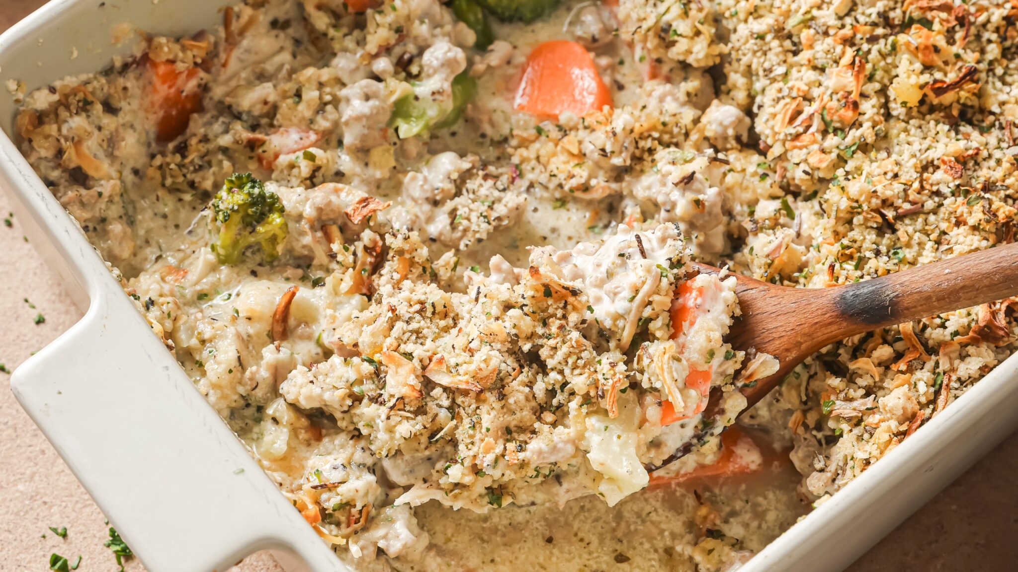 A close-up of a white baking dish filled with sausage stuffing casserole, showing a wooden spoon scooping a creamy mixture of cooked sausage, vegetables like carrots and broccoli, and a golden topping of herbs and crumbled stuffing.