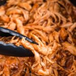 Close-up of tongs lifting saucy shredded barbecue chicken from a slow cooker.