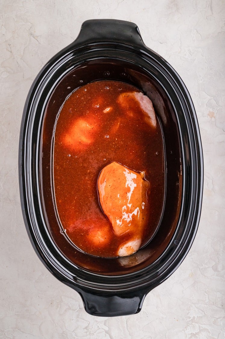 Raw chicken breasts in a slow cooker covered with a mixed barbecue sauce and seasoning liquid.
