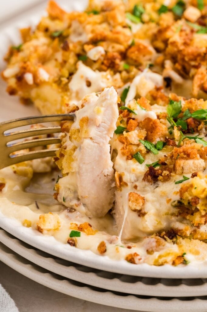 A fork cutting into creamy baked chicken topped with golden, crunchy stuffing and chopped herbs on a white plate.