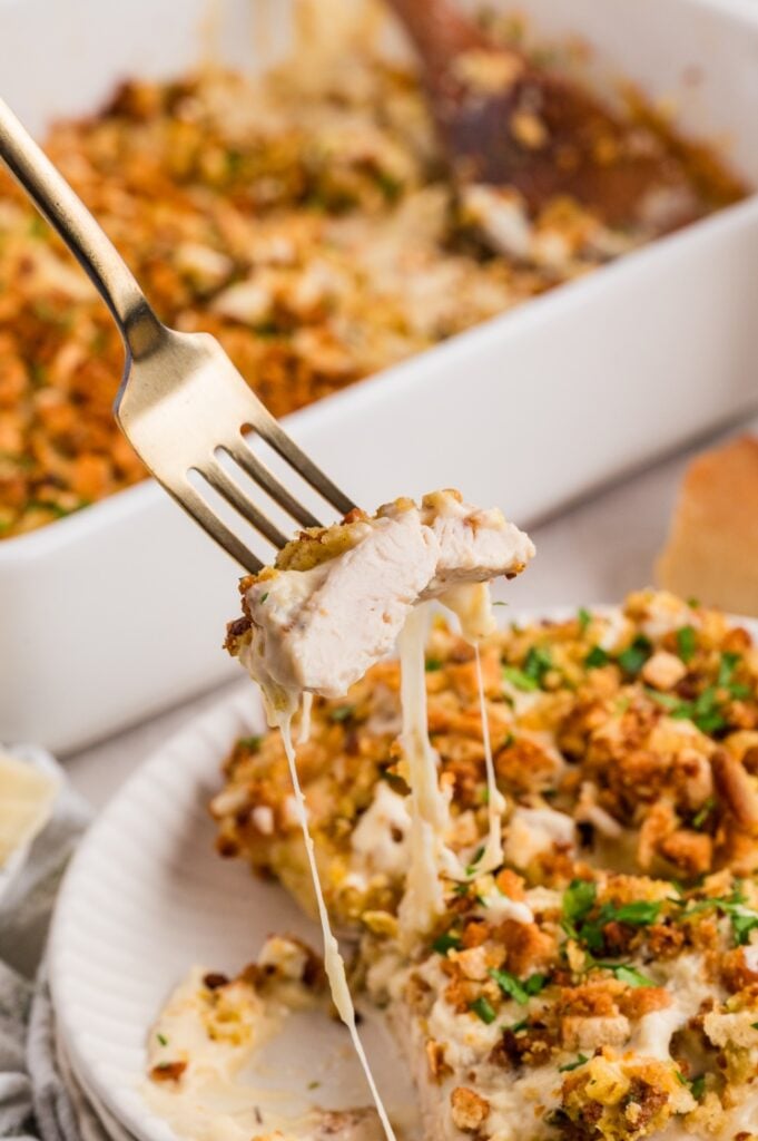 A fork holds a cheesy bite of baked chicken with a golden crumb topping, lifted from a plate. The plate and a baking dish full of the same dish are visible in the background.