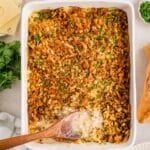 A baked casserole topped with golden, crispy crumbs and chopped herbs in a white dish, with a wooden spoon scooping out a creamy serving. Slices of bread, cheese, and fresh parsley are nearby on the table.