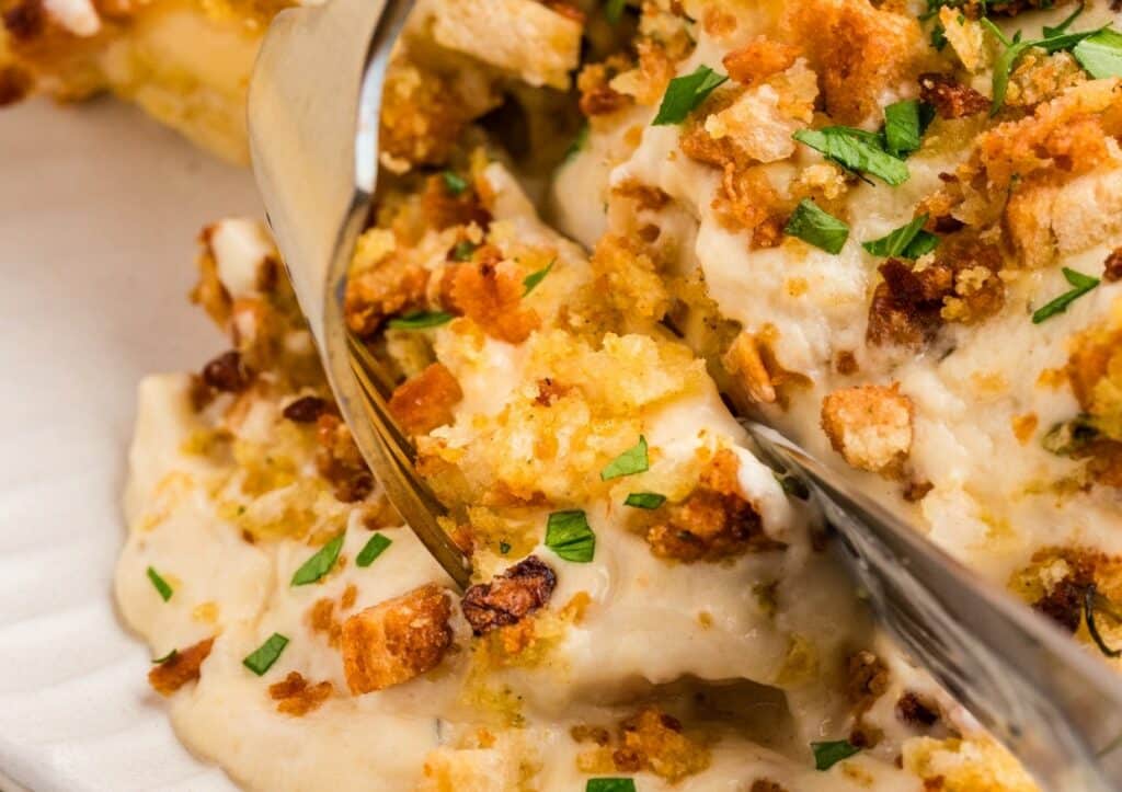 A close-up of a creamy casserole topped with golden brown breadcrumbs and chopped parsley, being served with a metal spatula.