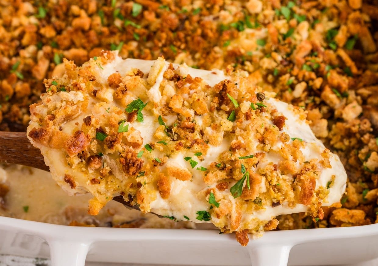 A wooden spoon scoops a cheesy, creamy casserole topped with golden, crispy stuffing and chopped parsley from a white baking dish.