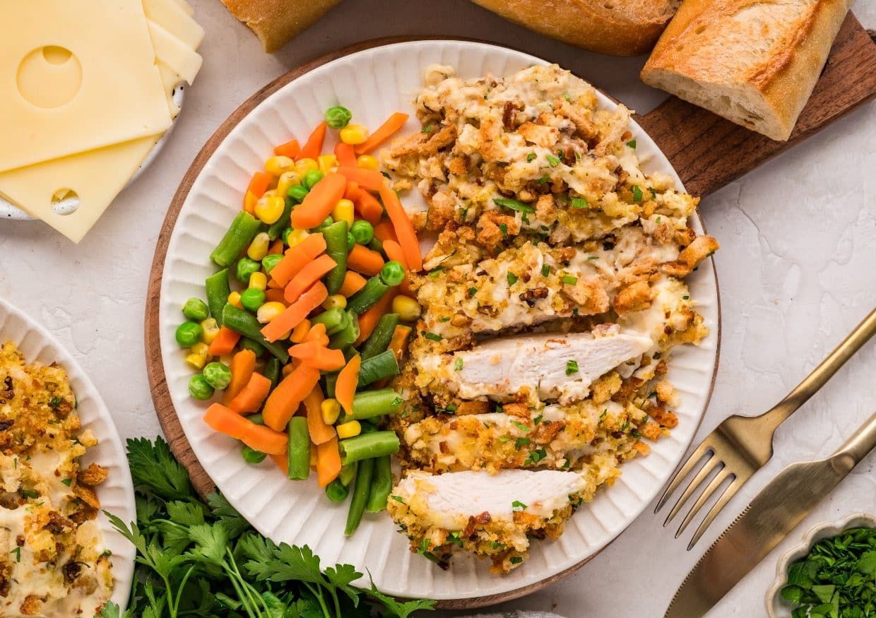 A plate with sliced chicken topped with stuffing, served with mixed vegetables (carrots, peas, green beans, and corn). Slices of baguette, Swiss cheese, and herbs are nearby. A fork and knife are beside the plate.
