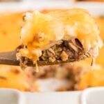 A close-up of a wooden spatula holding a slice of cheesy baked casserole with shredded meat, beans, and sauce, above a white baking dish.