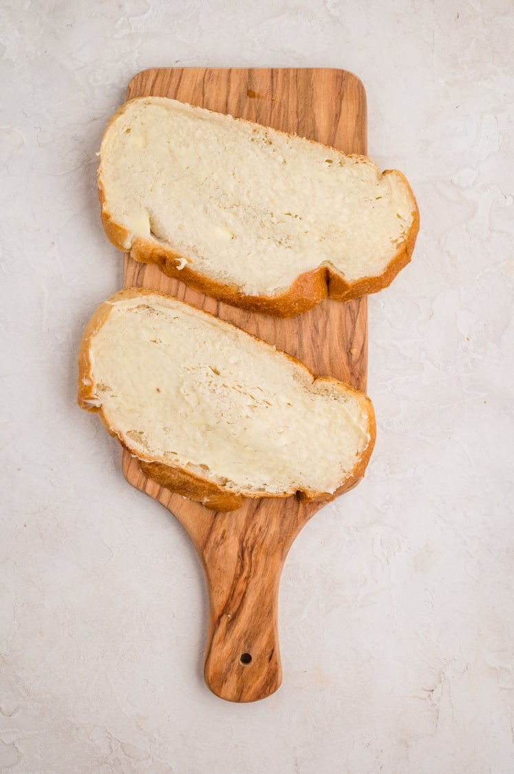 Two slices of bread with butter spread on top are placed on a wooden cutting board against a light, textured background.