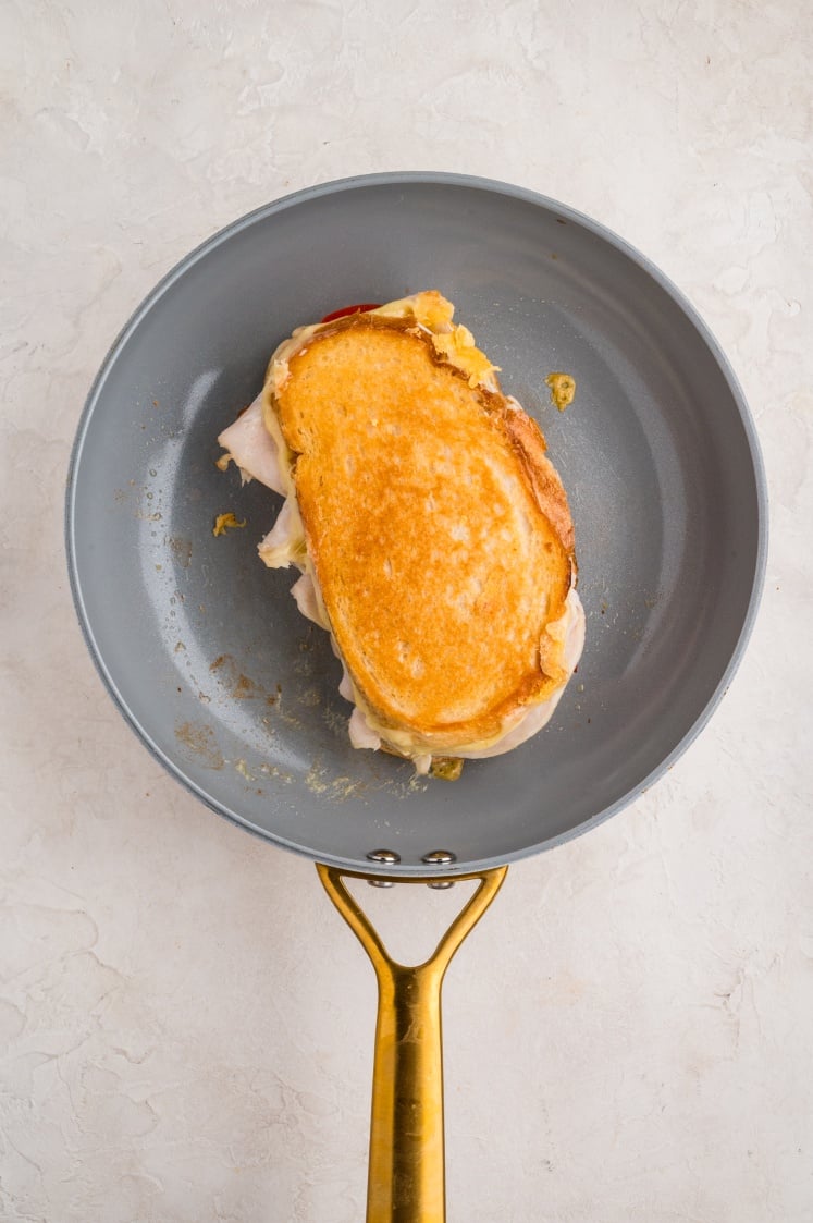 A golden-brown grilled cheese sandwich with turkey slices in a gray frying pan with a gold handle, set on a light-colored surface.