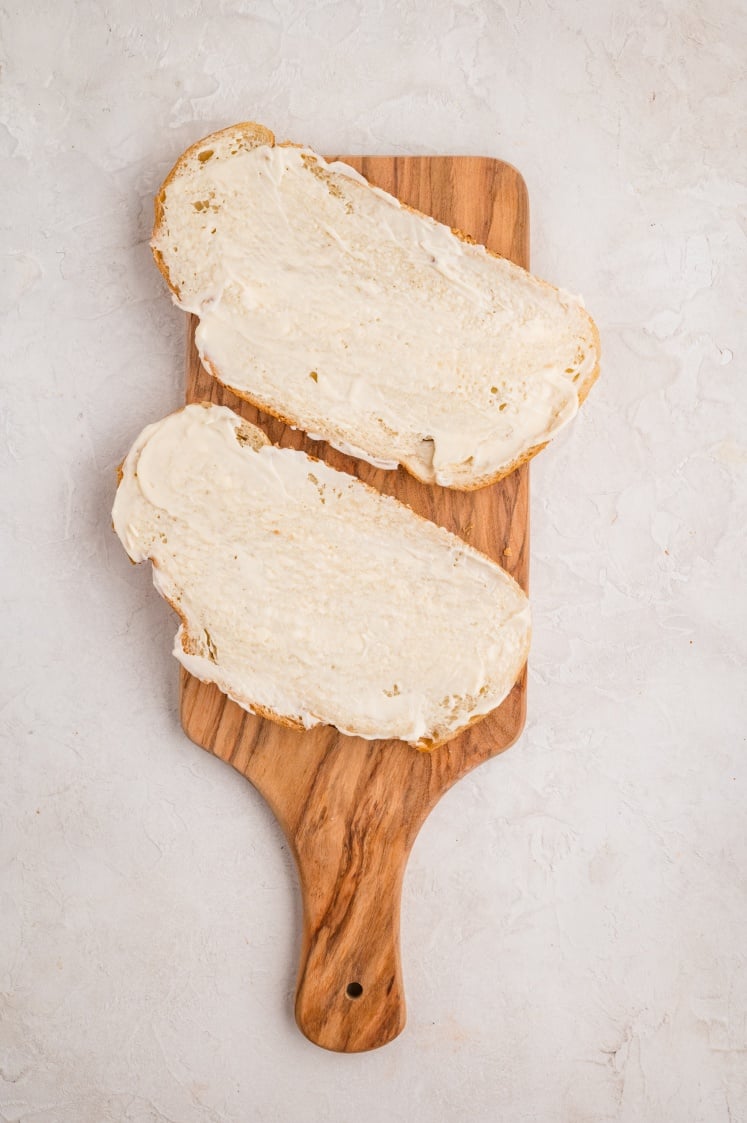 Two slices of bread with a layer of butter spread on them, placed on a wooden cutting board with a handle, set against a light, textured background.