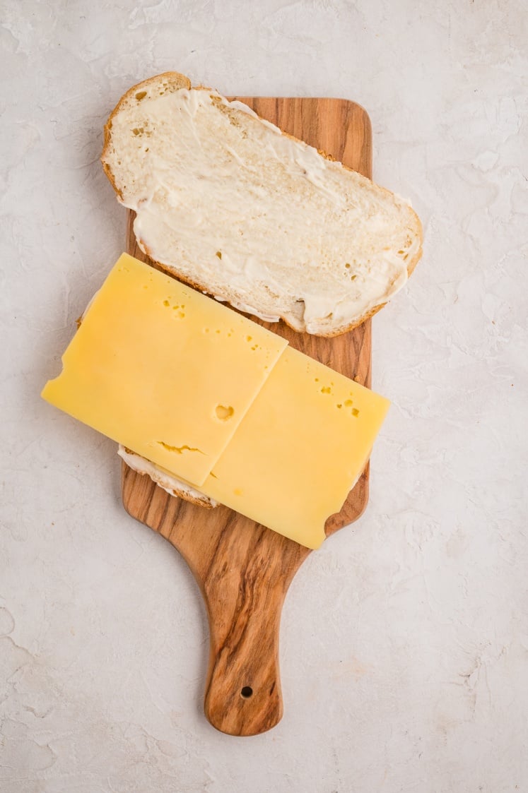 A wooden cutting board holds an open-faced sandwich with two slices of white bread, one spread with mayonnaise and the other topped with two slices of Swiss cheese.