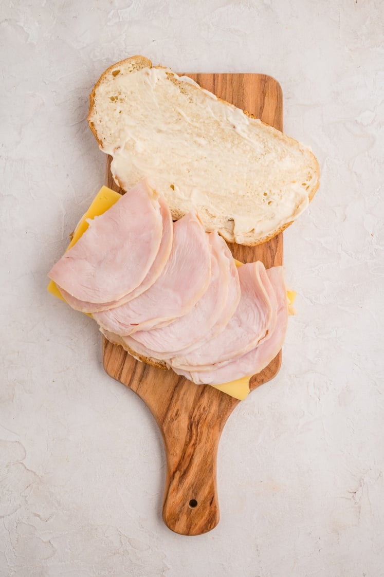 A wooden cutting board holds an open-faced sandwich with slices of ham and cheese on bread, and the top slice spread with mayonnaise, resting beside it.