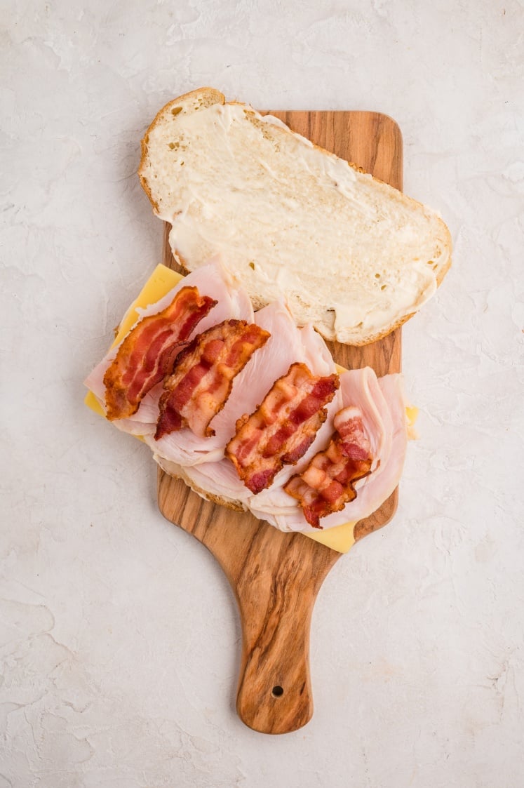 An open-faced sandwich on a wooden board, topped with slices of turkey, cheddar cheese, crispy bacon, and a slice of bread spread with mayonnaise. The sandwich is arranged on a light-colored surface.