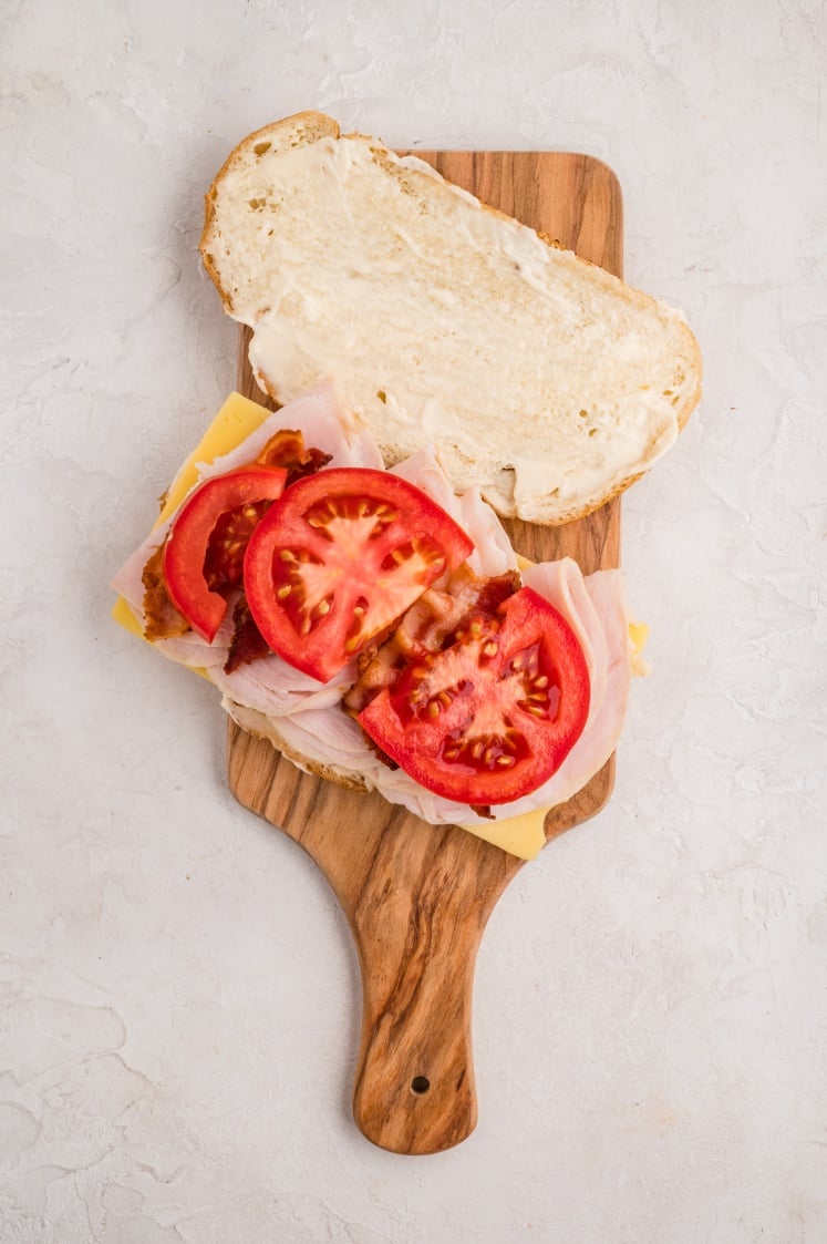 An open-faced sandwich on a wooden board, topped with sliced turkey, cheese, crispy bacon, and fresh tomato slices, with mayonnaise spread on the top slice of bread.