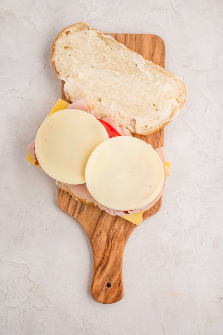 A sandwich on a wooden cutting board, with one slice of bread open to reveal layers of sliced cheese, deli meat, and mayonnaise on a light textured surface.
