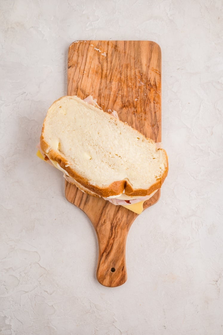 A sandwich with sliced bread, cheese, and deli meat sits on a wooden cutting board against a light, textured background.