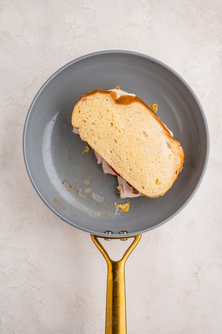 A sandwich with sliced bread and visible fillings is being toasted in a nonstick frying pan with a gold-colored handle, seen from above on a light surface.