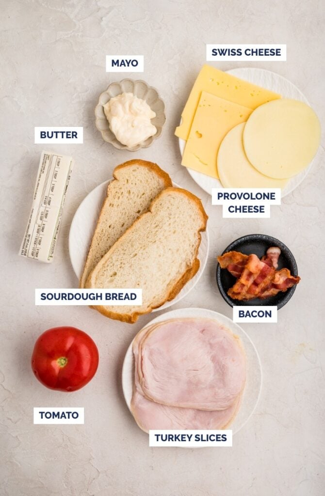 Ingredients for a sandwich are laid out on a white surface: a dish of mayo, slices of Swiss and provolone cheese, a stick of butter, two slices of sourdough bread, crispy bacon, a tomato, and turkey slices.