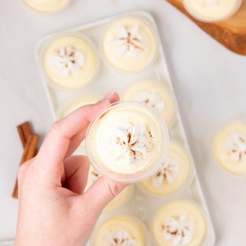 A hand holding a small cup of creamy dessert topped with whipped cream and a sprinkle of spice, with several similar desserts arranged on a tray beneath.