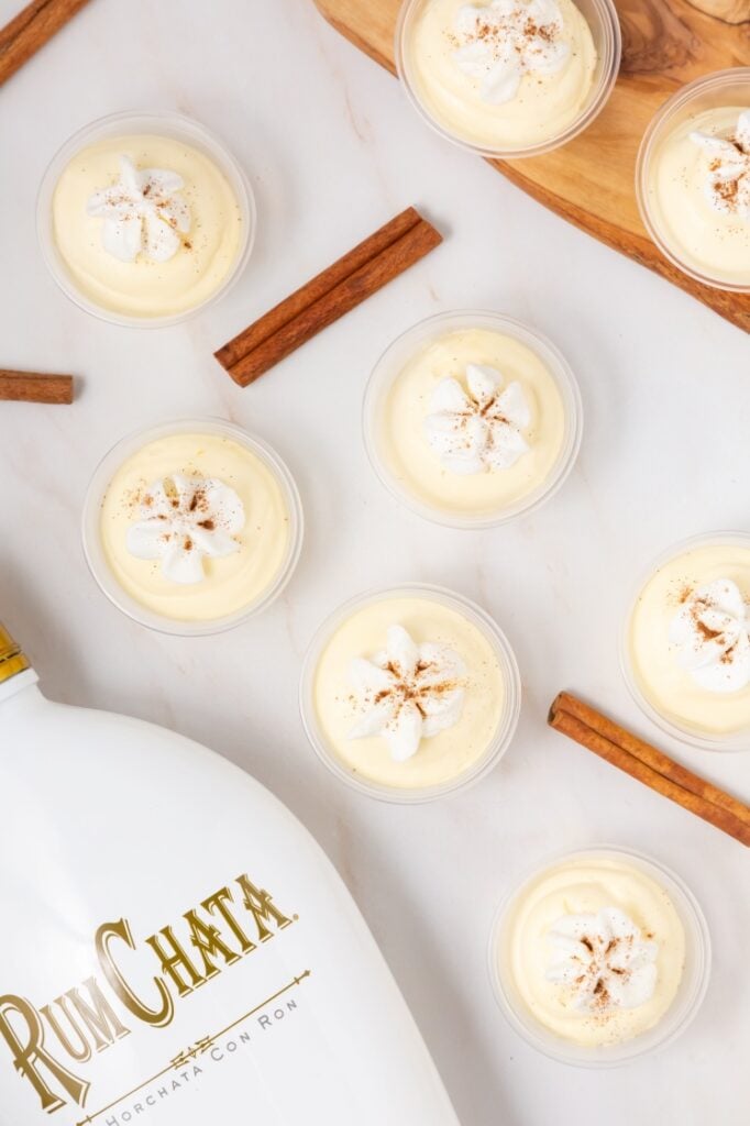 Small cups filled with creamy, light-yellow dessert topped with whipped cream and a sprinkle of cinnamon are arranged on a white surface with cinnamon sticks and a RumChata bottle nearby.