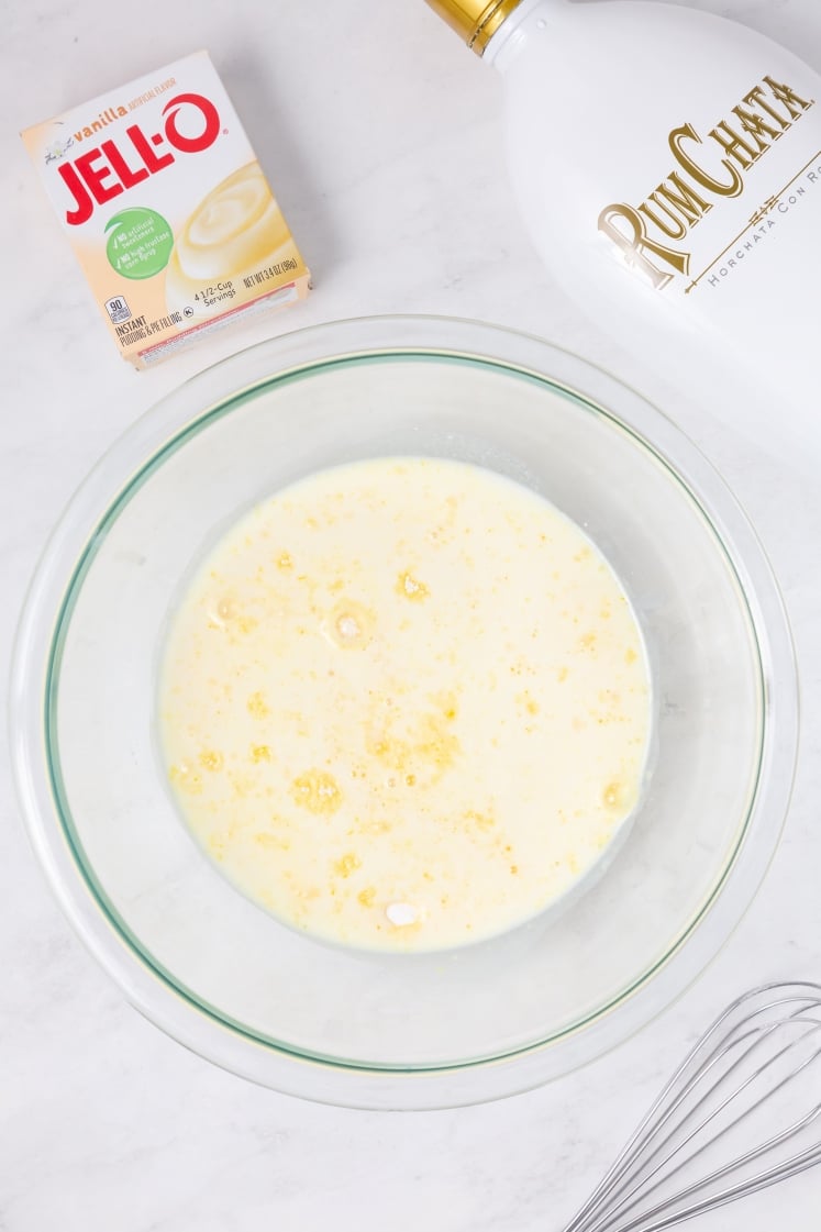 A glass mixing bowl filled with a creamy liquid mixture sits on a white surface, next to a box of Jell-O vanilla pudding mix, a bottle of RumChata, and a metal whisk.