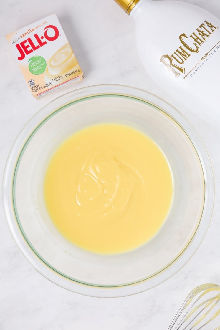 A glass bowl of yellow pudding mixture sits on a white surface, with a box of Jell-O vanilla pudding mix, a bottle of RumChata, and a whisk nearby.