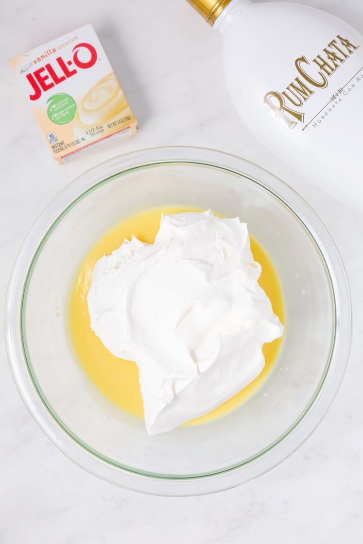 A glass bowl containing whipped topping and yellow pudding mixture sits on a white surface. Nearby are a box of Jell-O vanilla instant pudding and a white bottle labeled RumChata.