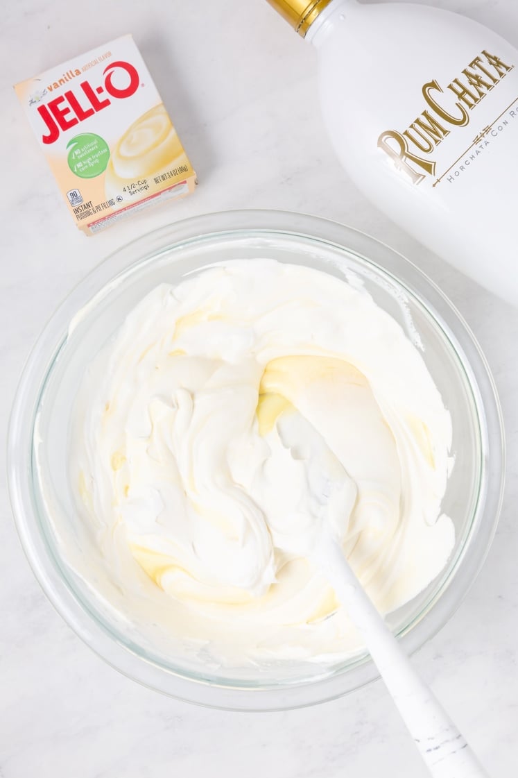 A bowl of whipped mixture with a spatula, next to a box of Jell-O vanilla pudding mix and a bottle of RumChata on a white surface.