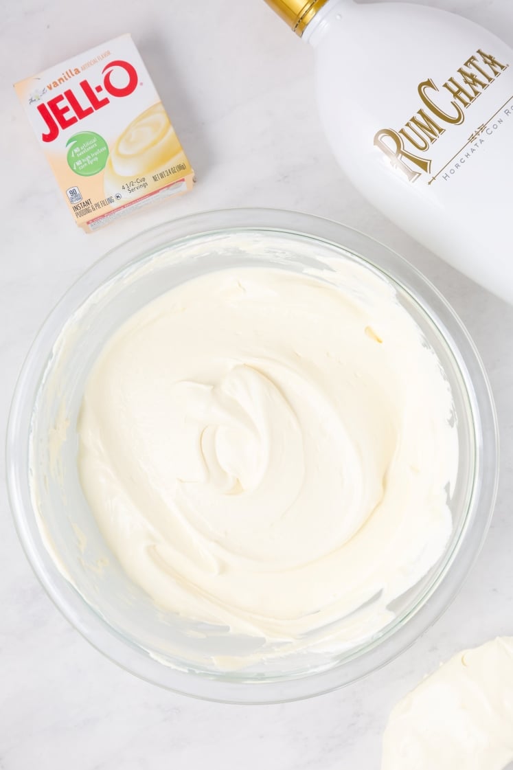 A glass bowl of creamy white mixture is on a marble surface, next to a box of Jello vanilla instant pudding and a bottle of RumChata liqueur.