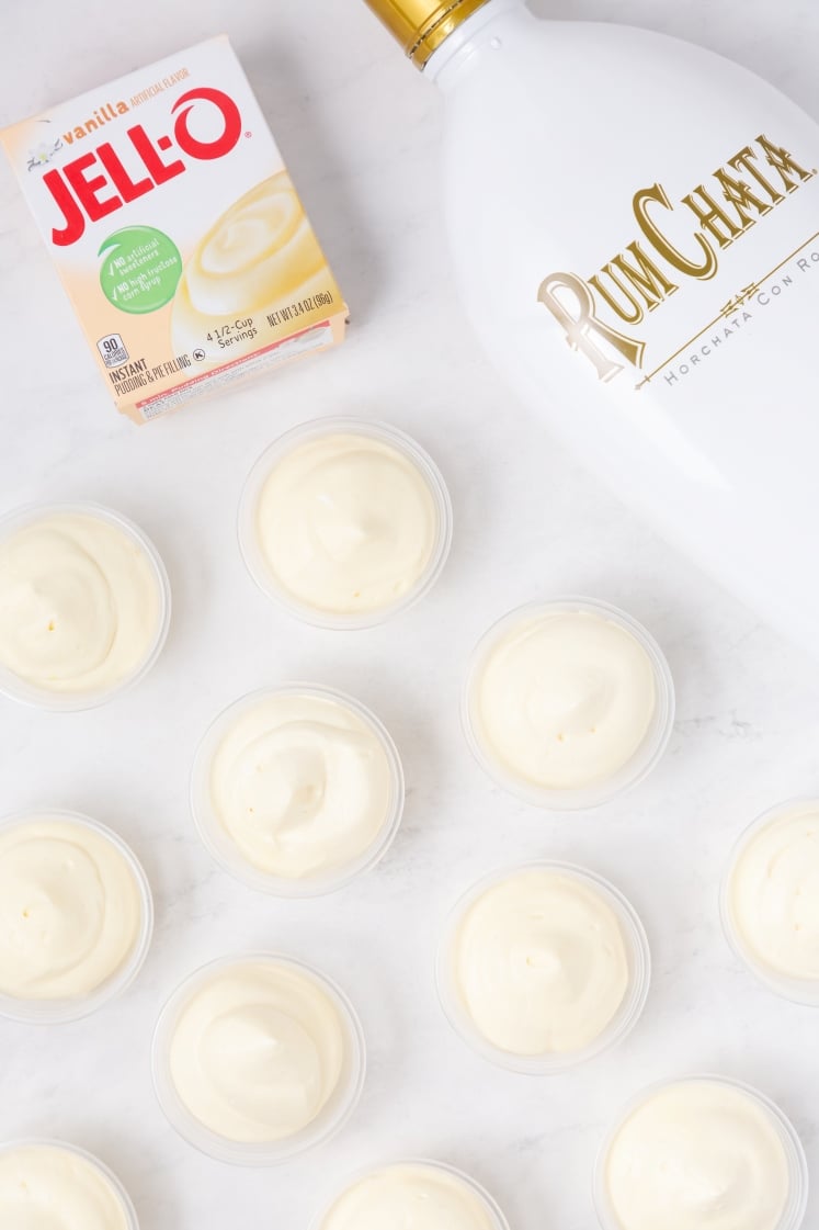 A bottle of RumChata, a box of Jell-O vanilla instant pudding, and several small cups filled with creamy white pudding are arranged on a white surface.
