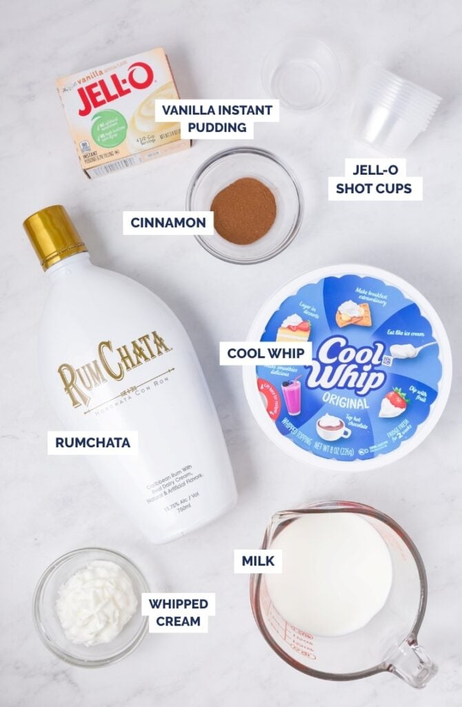 Ingredients for a dessert are arranged on a white surface: vanilla instant pudding, cinnamon, Jell-O shot cups, RumChata, Cool Whip, whipped cream, and milk, each labeled with text.
