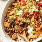 A white baking dish filled with taco casserole topped with Fritos, ground beef, corn, beans, shredded cheese, lettuce, diced tomatoes, green onions, and dollops of sour cream. A wooden spoon rests in the dish.