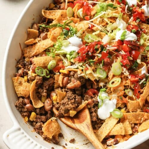 A white baking dish filled with taco casserole topped with Fritos, ground beef, corn, beans, shredded cheese, lettuce, diced tomatoes, green onions, and dollops of sour cream. A wooden spoon rests in the dish.
