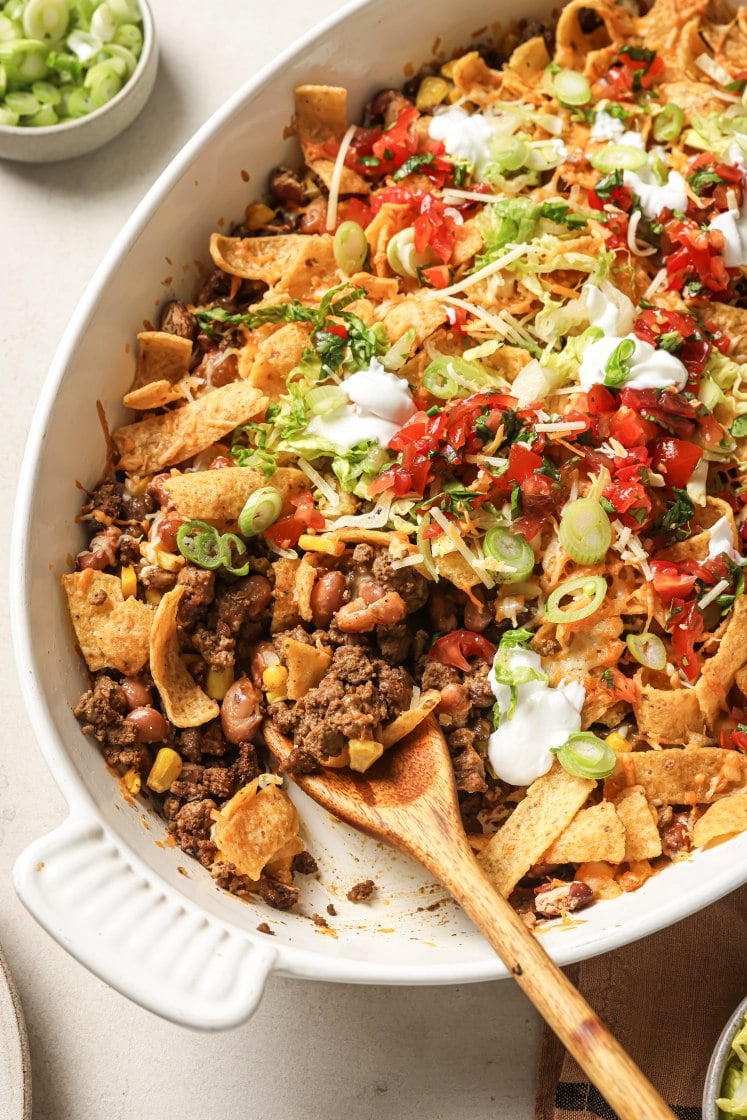 A white baking dish filled with taco casserole topped with Fritos, ground beef, corn, beans, shredded cheese, lettuce, diced tomatoes, green onions, and dollops of sour cream. A wooden spoon rests in the dish.
