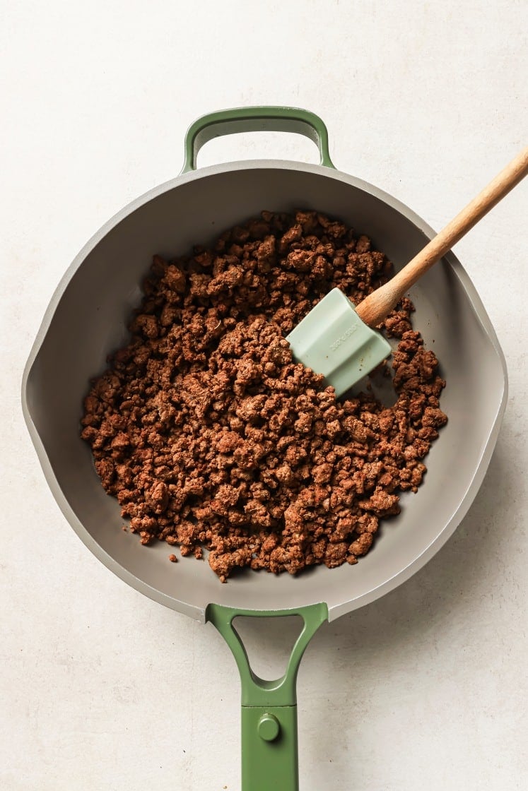 A green-handled skillet filled with cooked, browned ground beef sits on a light surface. A green spatula rests in the pan among the crumbled meat.