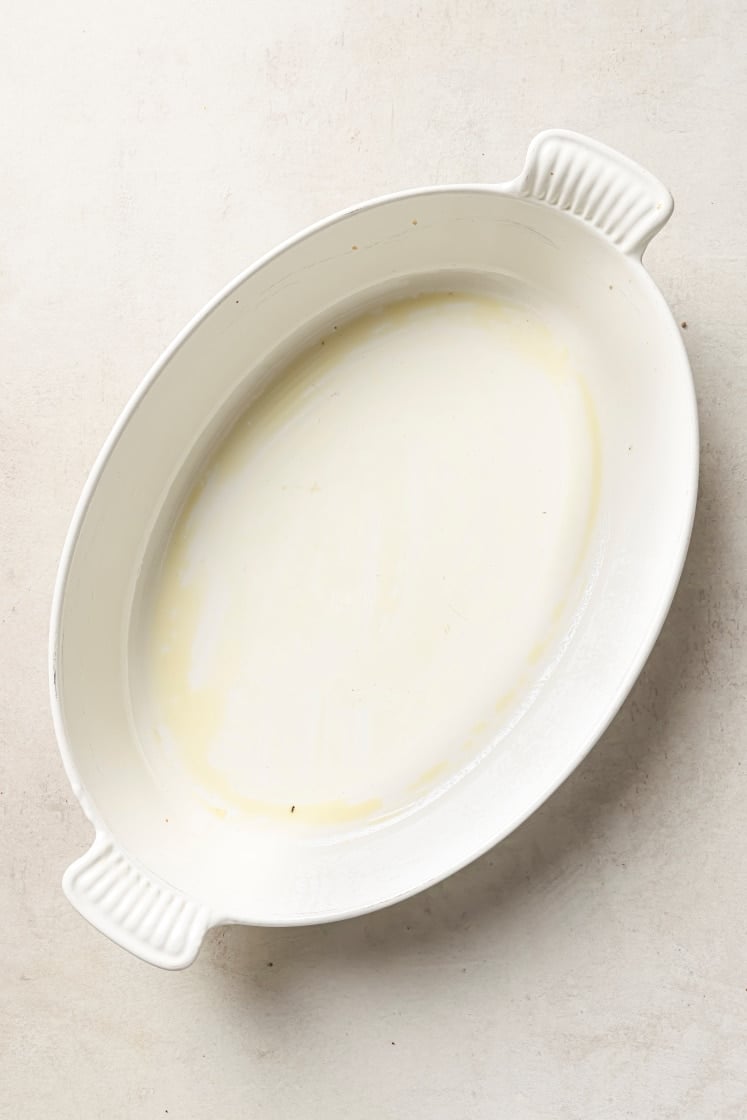 Oval white ceramic baking dish with ridged handles, lightly greased with a thin layer of oil or butter, on a light-colored surface.