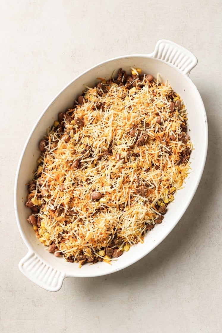 An oval white baking dish filled with a bean casserole topped with shredded cheese and crispy bits, on a light-colored surface.