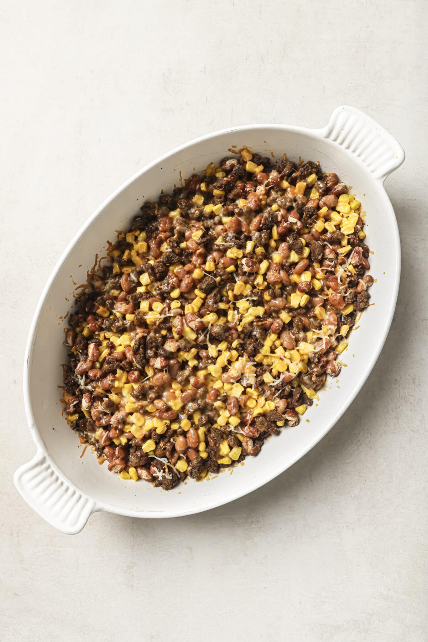 An oval white baking dish filled with a mixture of ground beef, corn, beans, and shredded cheese, placed on a light-colored surface.