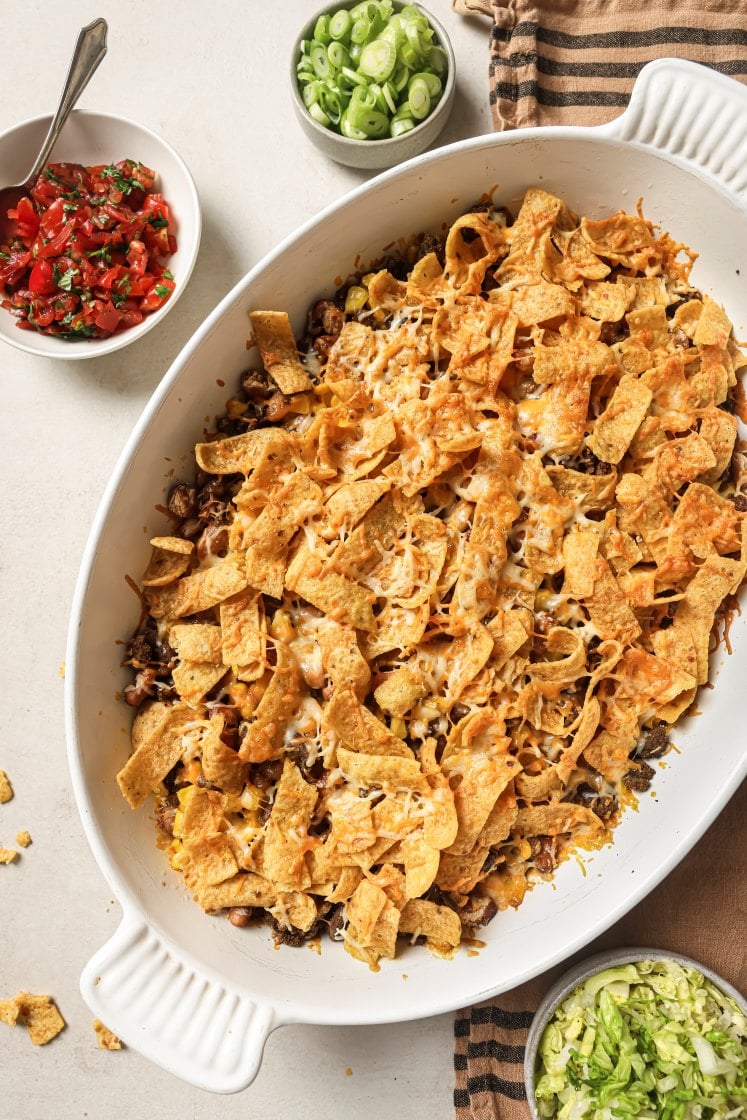 A white casserole dish filled with a baked Walking Taco Bake topped with melted cheese and corn chips. Nearby are bowls of chopped green onions, salsa, and shredded lettuce on a light surface with a striped towel.