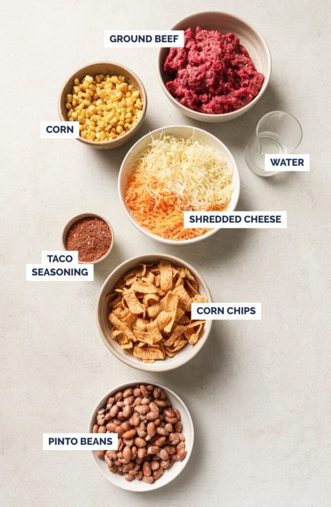 Five bowls containing labeled ingredients of walking taco bake: ground beef, corn, shredded cheese, corn chips, and pinto beans, along with a glass of water and a small bowl of taco seasoning, all arranged on a light surface.