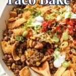 A casserole dish filled with walking taco bake, featuring ground beef, corn chips, beans, cheese, tomatoes, green onions, and dollops of sour cream. A wooden spoon rests in the dish. Text reads "Walking Taco Bake.