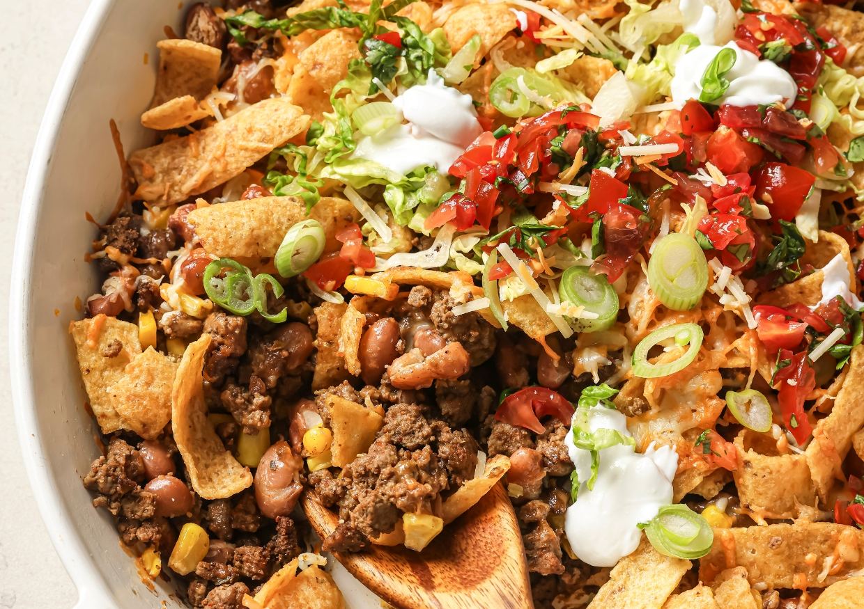 A casserole dish of walking taco bake topped with toppings and a wooden spoon.