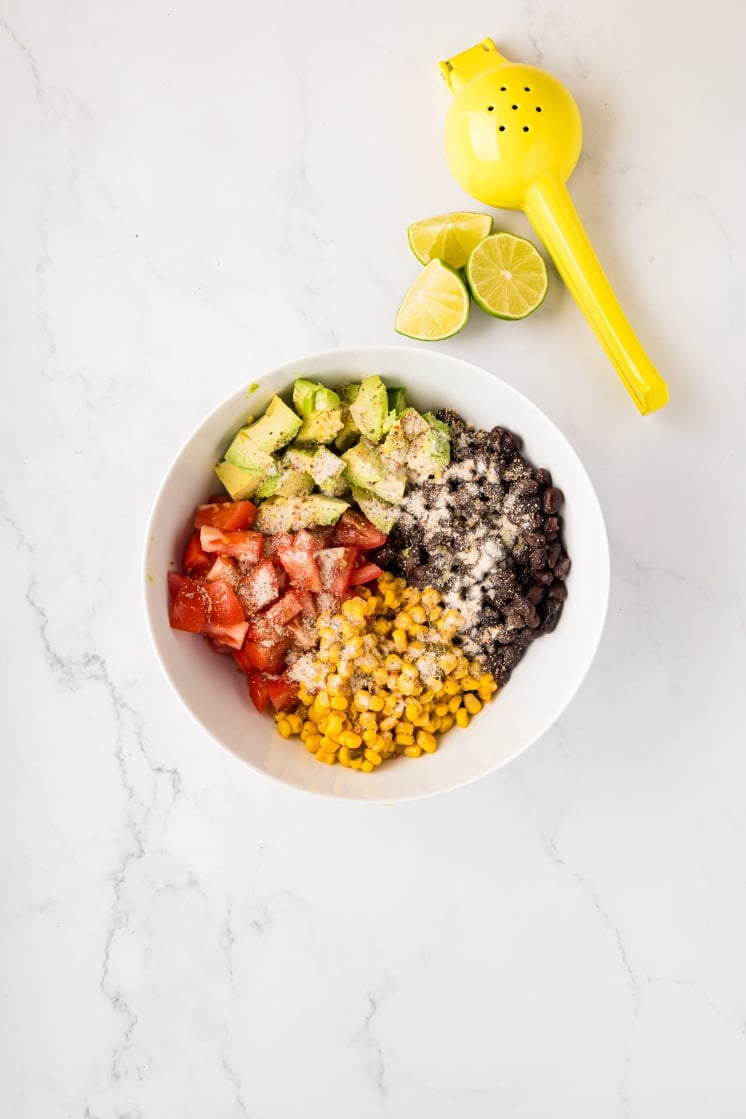 A white bowl filled with diced avocado, tomatoes, corn and grated cheese, arranged separately. Beside the bowl are lime halves and a yellow citrus squeezer on a white marble surface.