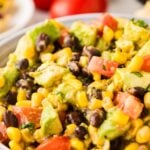 A close-up of a bowl filled with a colorful salad made of black beans, corn, diced tomatoes, avocado, and herbs. The background shows blurred tomatoes and limes.
