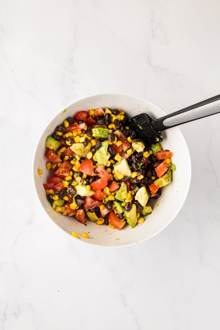 A white bowl filled with a colorful salad of diced tomatoes, avocado, black beans, and corn, with a black spatula resting inside, on a white marble surface.