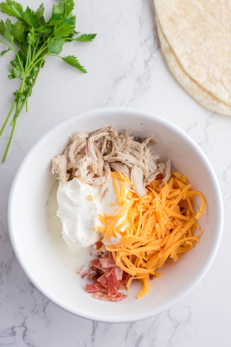 Bowl filled with ingredients for crack chicken pinwheels, including shredded chicken, cream cheese, cheddar cheese, bacon, and ranch dressing, with tortillas and parsley nearby.
