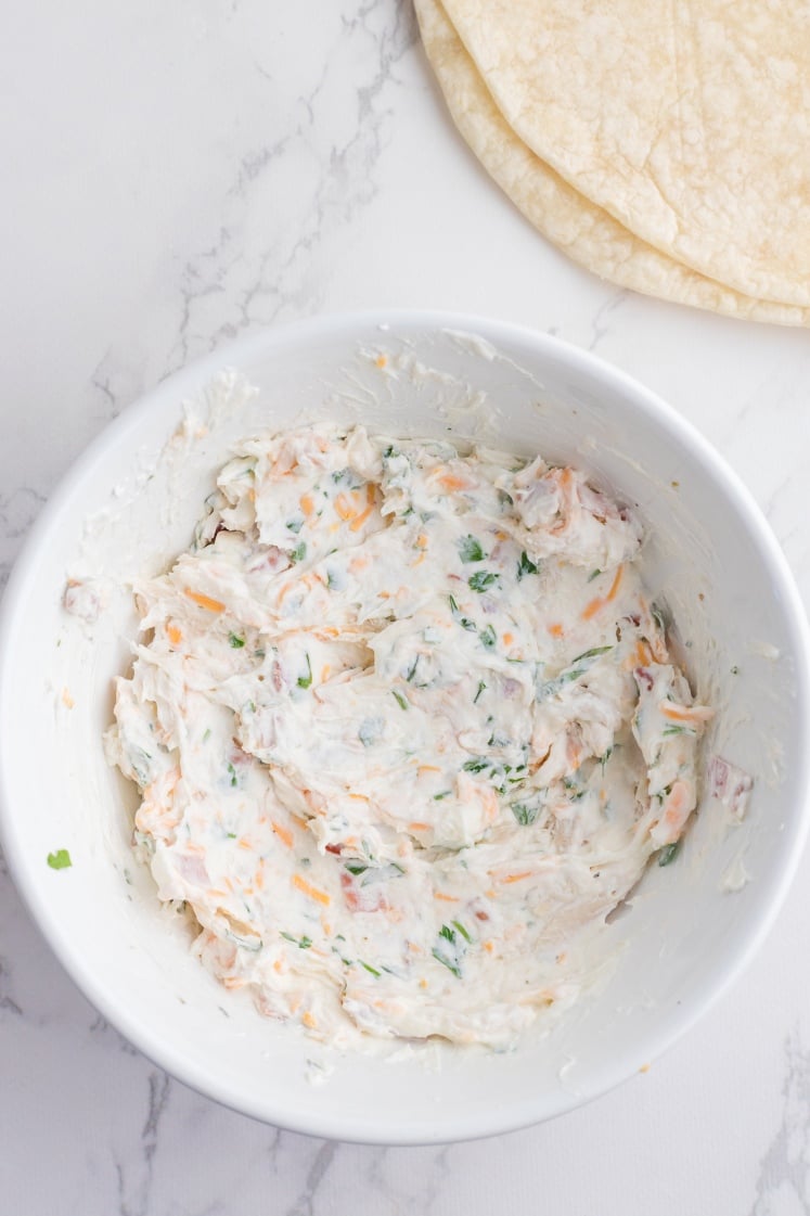 Bowl of mixed crack chicken pinwheel filling made with cream cheese, cheddar, bacon, chicken, and parsley, with tortillas ready for assembling.