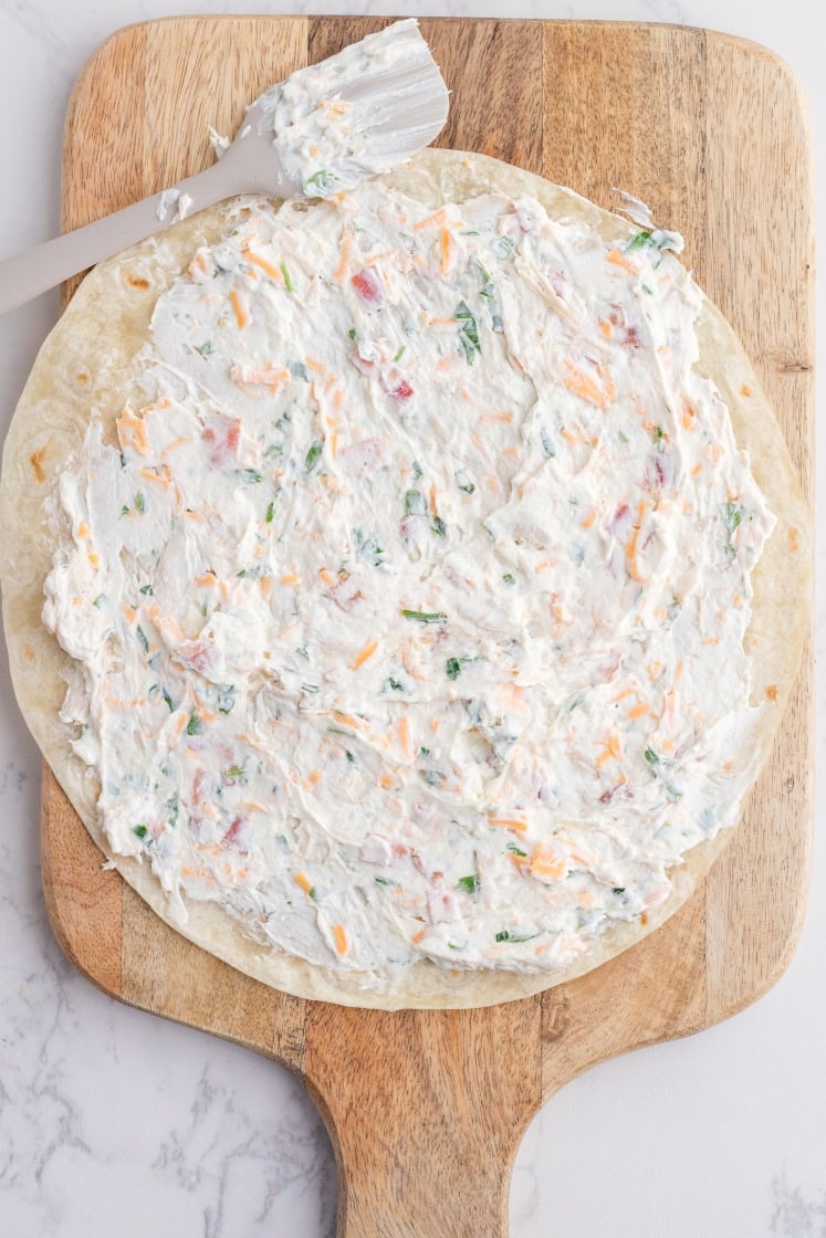 Tortilla spread with crack chicken pinwheel filling made of cream cheese, cheddar, bacon, chicken, and herbs on a wooden board.