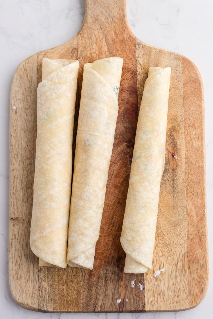 Three tortillas rolled tightly with crack chicken pinwheel filling on a wooden cutting board.