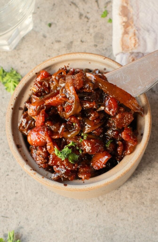 A ceramic bowl filled with chunky bacon jam topped with herbs, with a knife resting inside, placed on a stone surface.